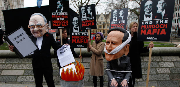 A protester in London, dressed as a caricature of News Corp. Chairman Rupert Murdoch, burns a government report on media abuses while another wearing a mask depicting Prime Minister David Cameron sits tied to a chair, November 29, 2012. (AP/Sang Tan)