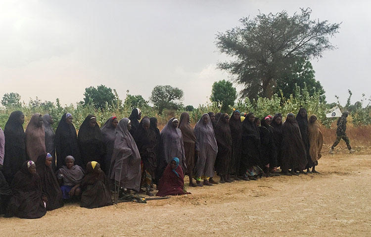 A man carrying a flag for the militant group Boko Haram walks past a group of 82 Chibok girls, who were held captive for three years by the militants, as the girls wait to be released in exchange for several militant commanders, near Kumshe, Nigeria on May 6, 2017. Nigeria's domestic intelligence agency has detained journalist Tony Ezimakor fin relation to an article he wrote about the release of the Nigerian schoolgirls, according to media reports. (Reuters/Zanah Mustapha)
