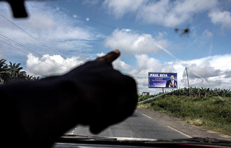 A taxi driver points towards an electoral poster for Cameroonian President Paul Biya as the road crosses into the majority English-speaking South West region of Cameroon, in Buea, on October 3, 2018. A journalist was detained October 23 after publishing articles criticizing the government's handling of grievances of Anglophone Cameroonians. (AFP/Marco Longari)
