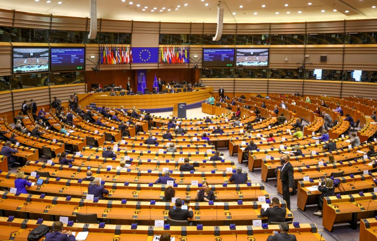 Lawmakers are shown seated at desks in rows facing a podium and EU flags in a large parliamentary building.