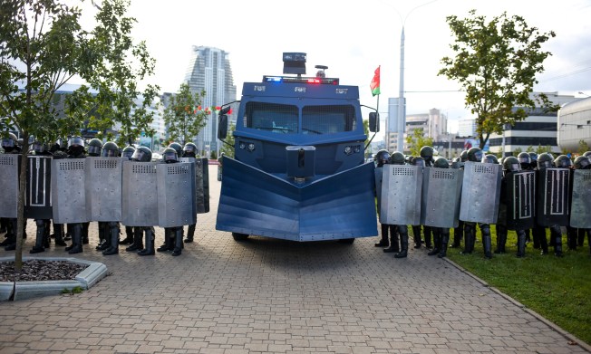 A large vehicle flanked by police officers holding shields blocks a city street.