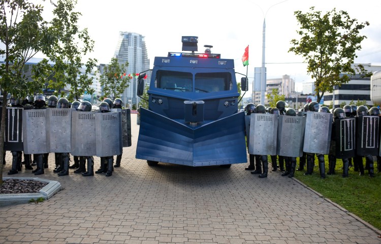 A large vehicle flanked by police officers holding shields blocks a city street.