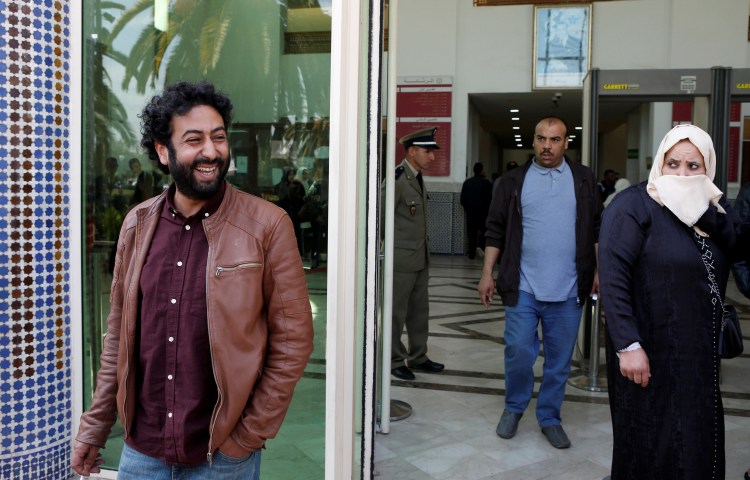 A man smiles for photographers in front of a doorway as other people exiting the same building look on.