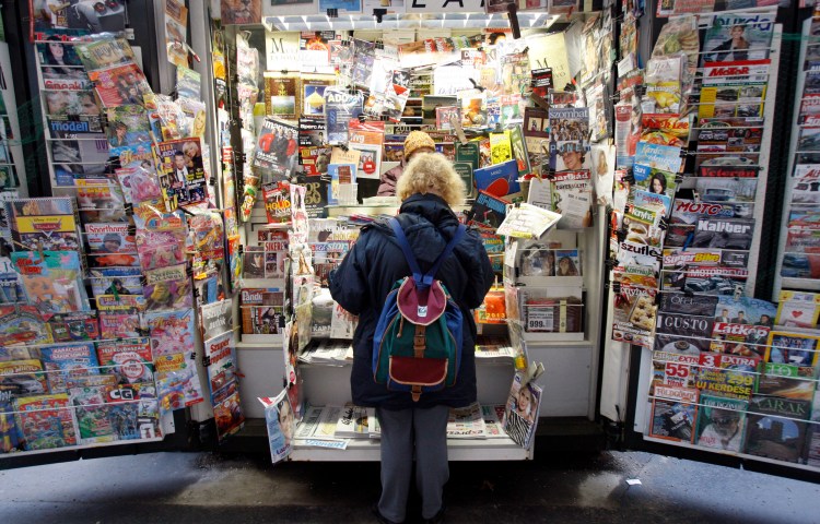 A woman facing away from the camera looks at a large display of magazines and newspapers.