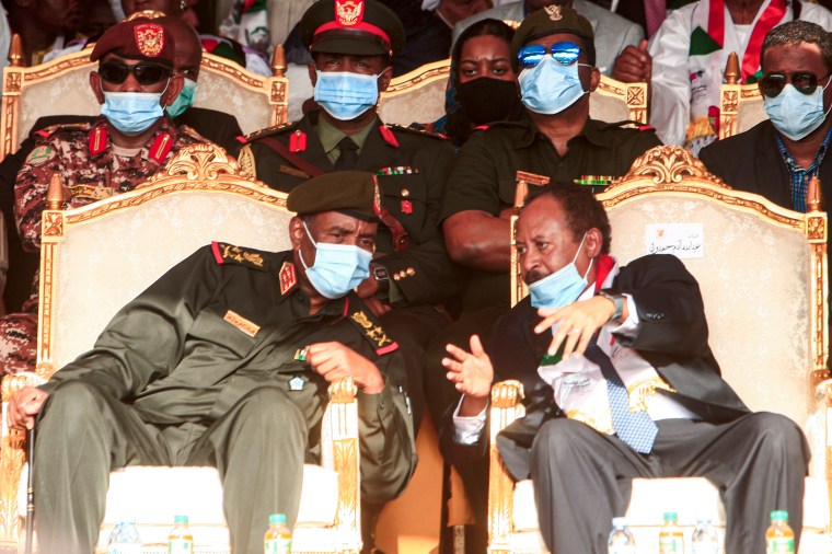 A man in military uniform and a coronavirus mask seated in an elaborate chair leans over to talk with a man in a suit and a coronavirus mask in the neighboring chair.