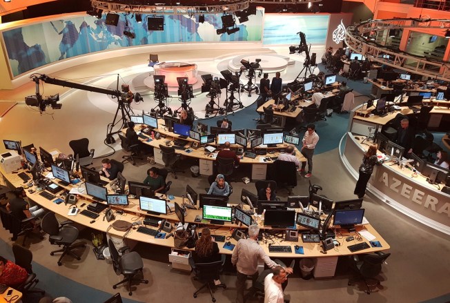 Journalists are shown working at their desks behind the scenes of a TV news studio.