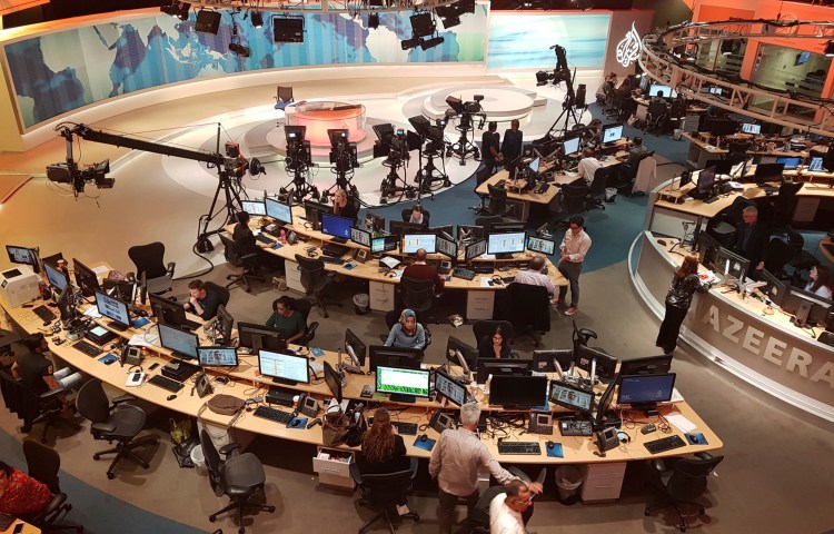 Journalists are shown working at their desks behind the scenes of a TV news studio.