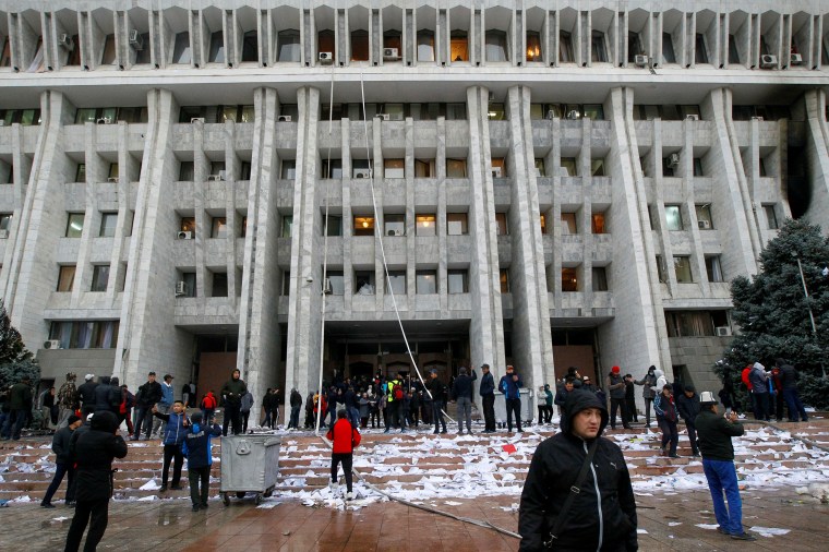 Figures are seen in the foreground of a large government building surrounded by trash and discarded paper.