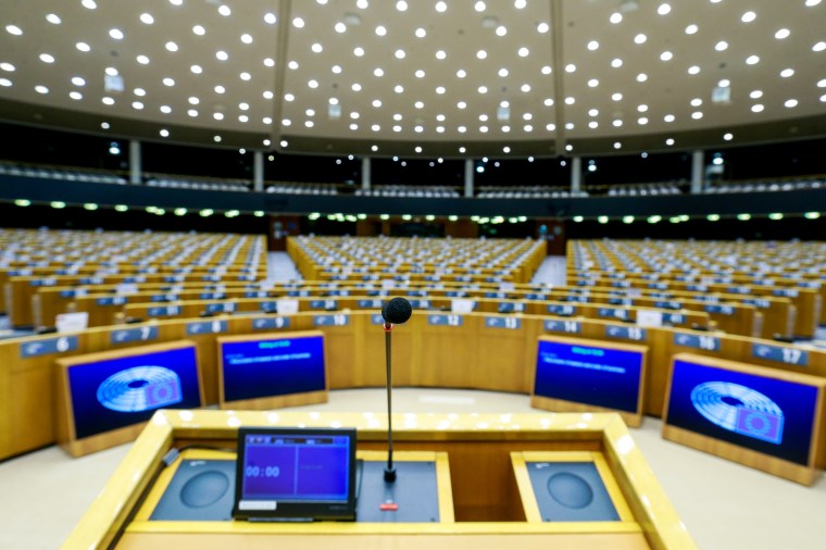 A lectern is shown against a backdrop of empty desks set up for a parliamentary meeting.