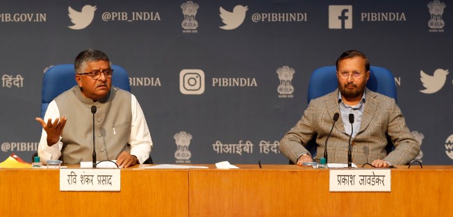 Two men sit at a podium against a backdrop illustrated with logos of social media companies.