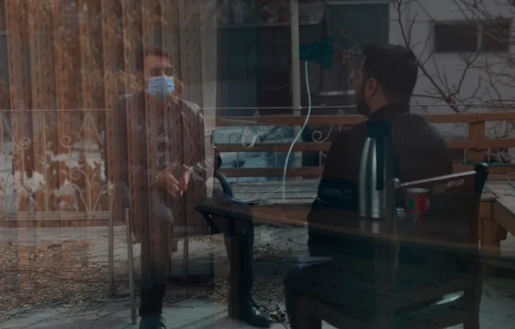 Two men partially obscured by a reflection in a window are shown talking, one wearing sunglasses and face mask.