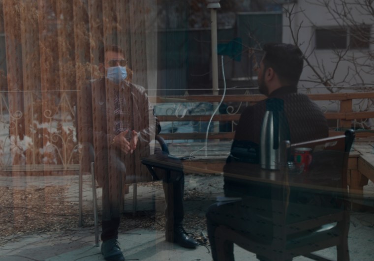 Two men partially obscured by a reflection in a window are shown talking, one wearing sunglasses and face mask.