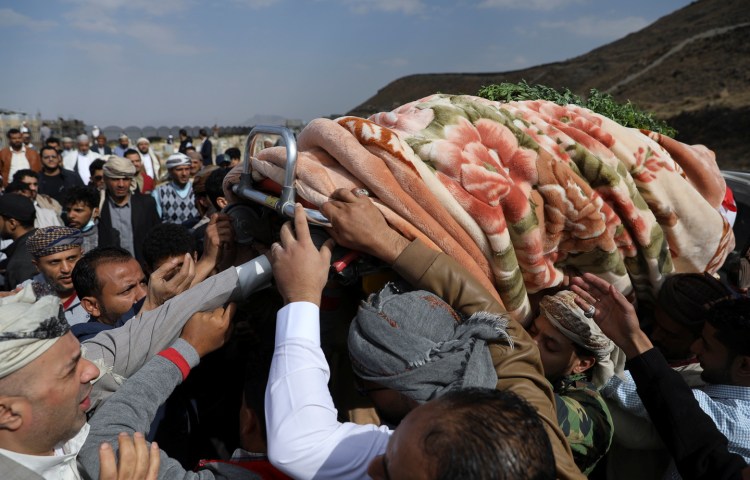 A crowd of men reach up to support a coffin draped in a blanket.