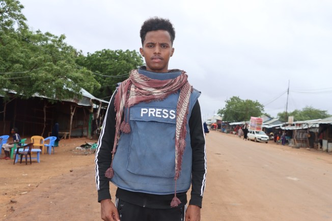 Mohamed Ibrahim Osman Bulbul of privately owned broadcaster Kaab TV stands on the side of a road, wearing a blue flak jacket marked 'Press'.