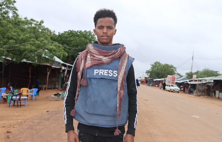 Mohamed Ibrahim Osman Bulbul of privately owned broadcaster Kaab TV stands on the side of a road, wearing a blue flak jacket marked 'Press'.