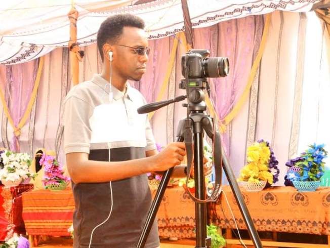 Abdifatah Yusuf Beereed of Goobjoog TV stands behind a camera on a tripod, filming.