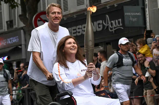 AFP video journalist Dylan Collins pushes the wheelchair of AFP photojournalist Christina Assi as she carries the Olympic flame on July 21 during the Olympic Torch Relay near Paris. Assi and Collins were injured in an attack by an Israeli tank on a group of journalists in southern Lebanon on October 13, 2023, that killed Reuters video journalist Issam Abdallah. (Photo: AFP/Mauro Pimentel)