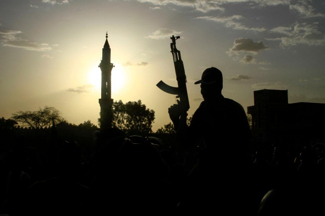 A fighter holds up a gun backdropped by the minaret of a mosque and the sun.