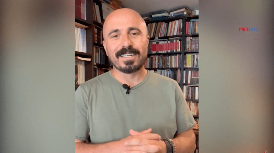 A man talks to a camera in front of a bookcase.