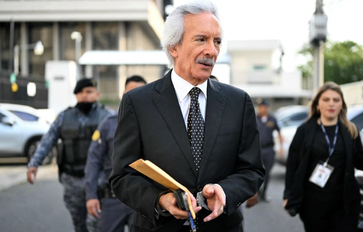 A handcuffed man in a suit walks carrying folders.