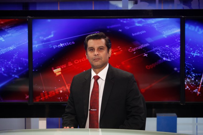 A man in a suit stands before a news broadcast background.