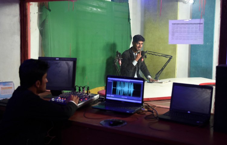 A man speaks on the radio while another man watches from the sound booth.