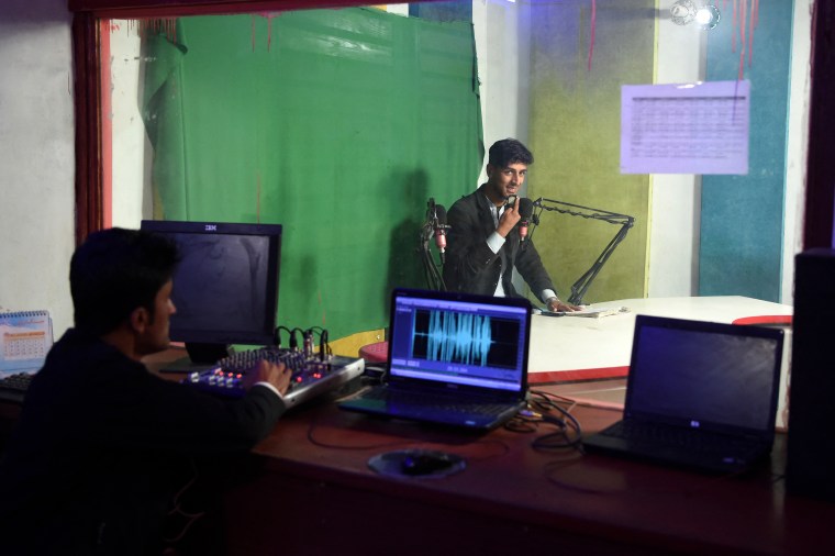 A man speaks on the radio while another man watches from the sound booth.