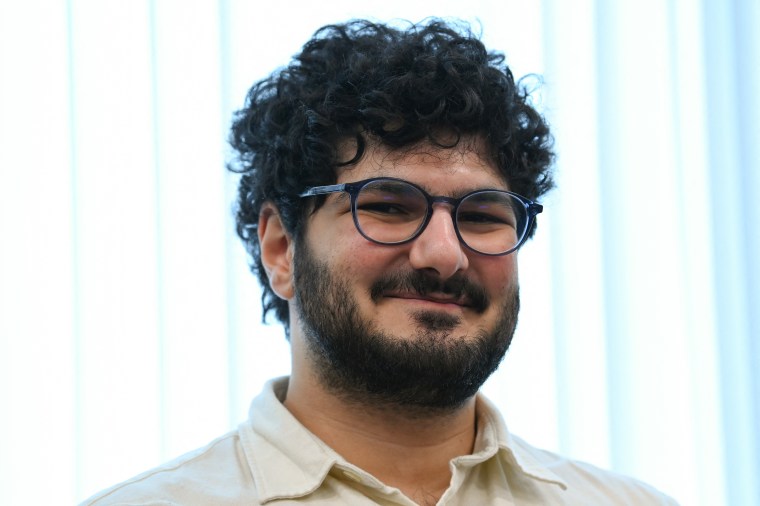 A man with curly hair and glasses smiles at the camera.