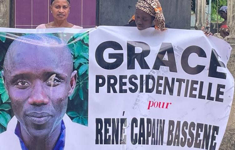 René Capain Bassène's wife, Odette Victorine Coly, holds a banner requesting a presidential pardon for the journalist during President Bassirou Diomaye Diakhar Faye's visit to Ziguinchor in December 2025. The banner reads, "Presidential pardon for René Capain Bassène."
