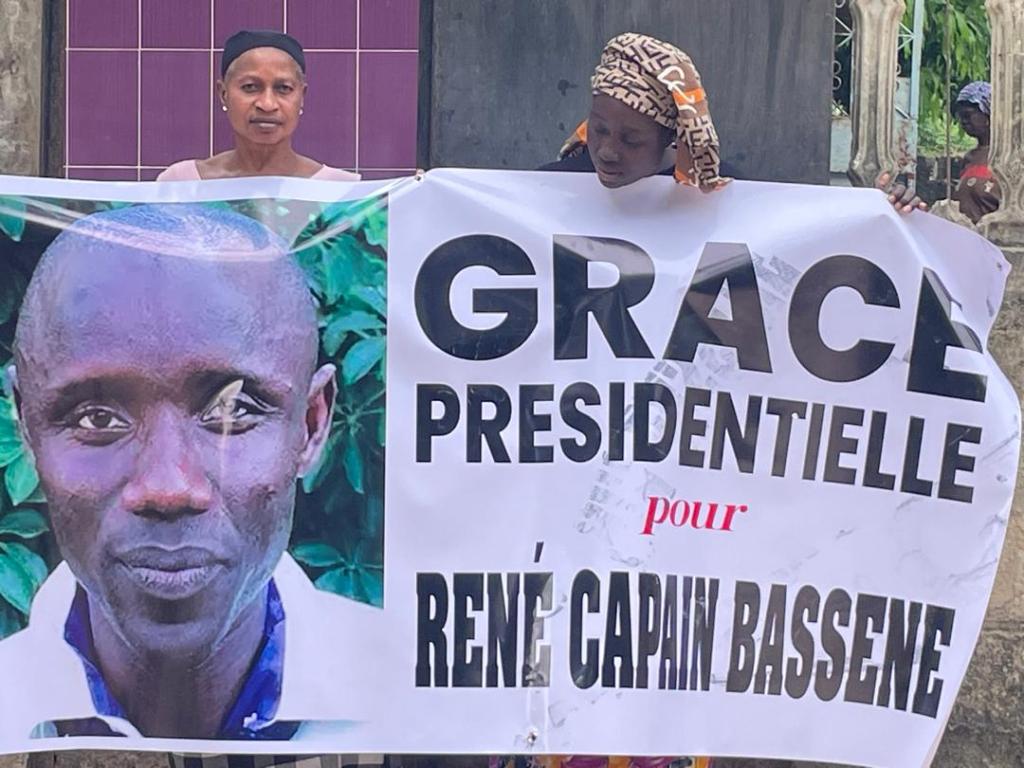 L’épouse de René Capain Bassène, Odette Victorine Coly, tient une banderole demandant une grâce présidentielle pour le journaliste lors de la visite du président Bassirou Diomaye Diakhar Faye à Ziguinchor en décembre 2025. La banderole indique : « Grâce présidentielle pour René Capain Bassène. »