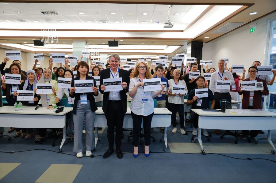 A group of people stand in a room holding up signs that say "# Free The Press" towards the camera.